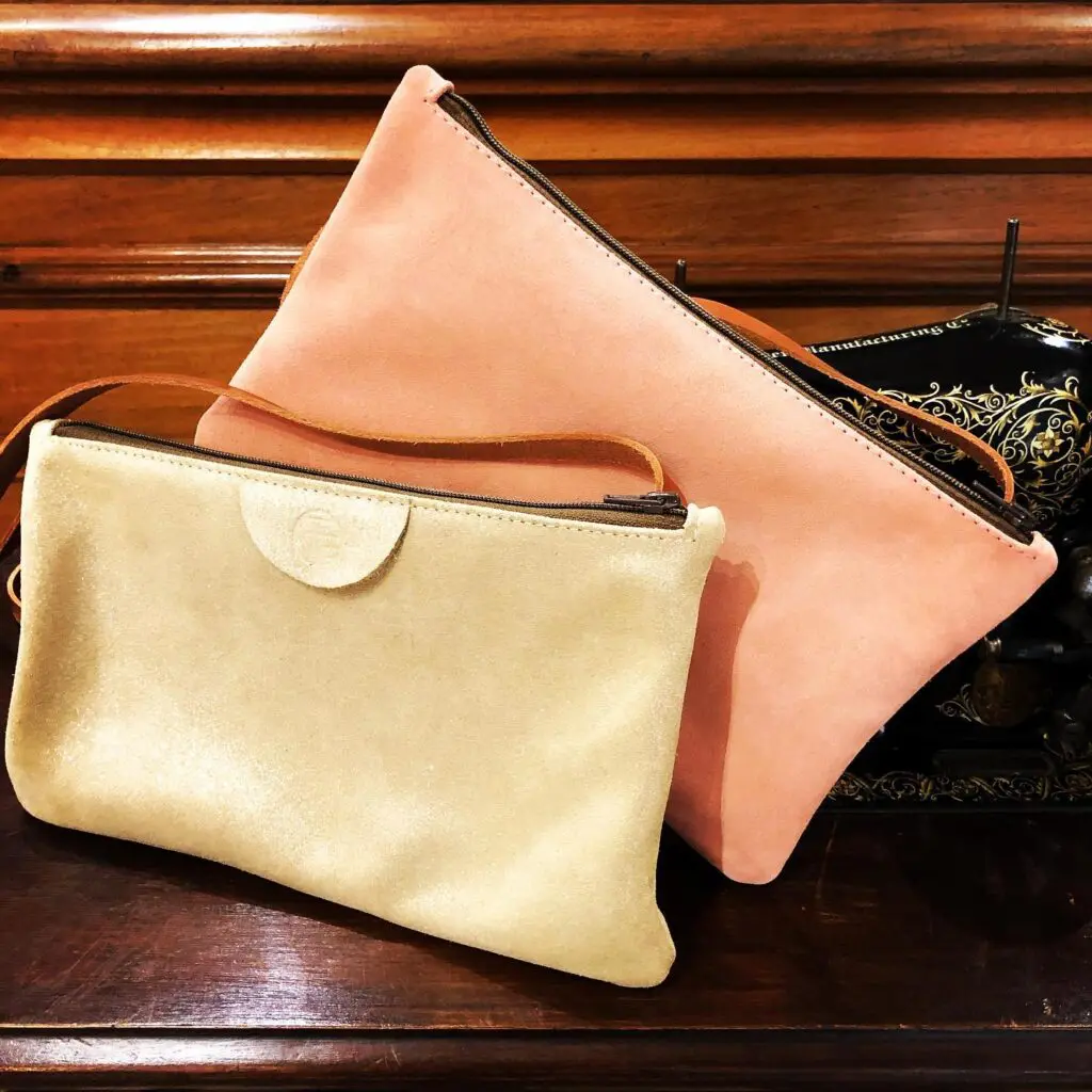 Deux pochettes en cuir, une beige et une rose, posées sur une surface en bois avec une machine à coudre en arrière-plan.