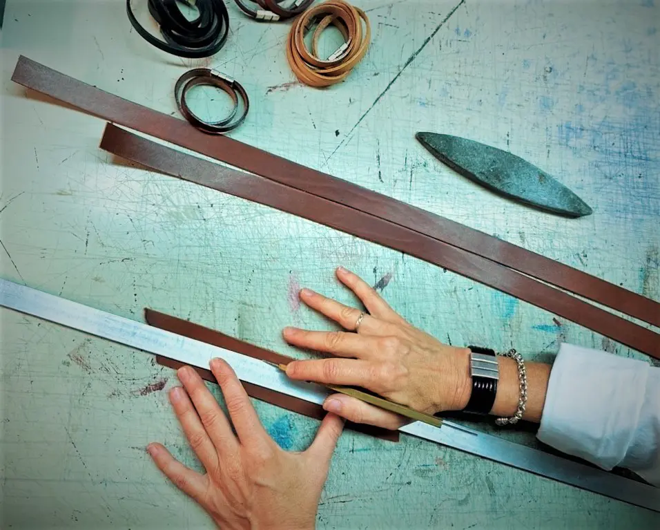 Mains mesurant des bandes de cuir sur une table avec des outils de maroquinerie.