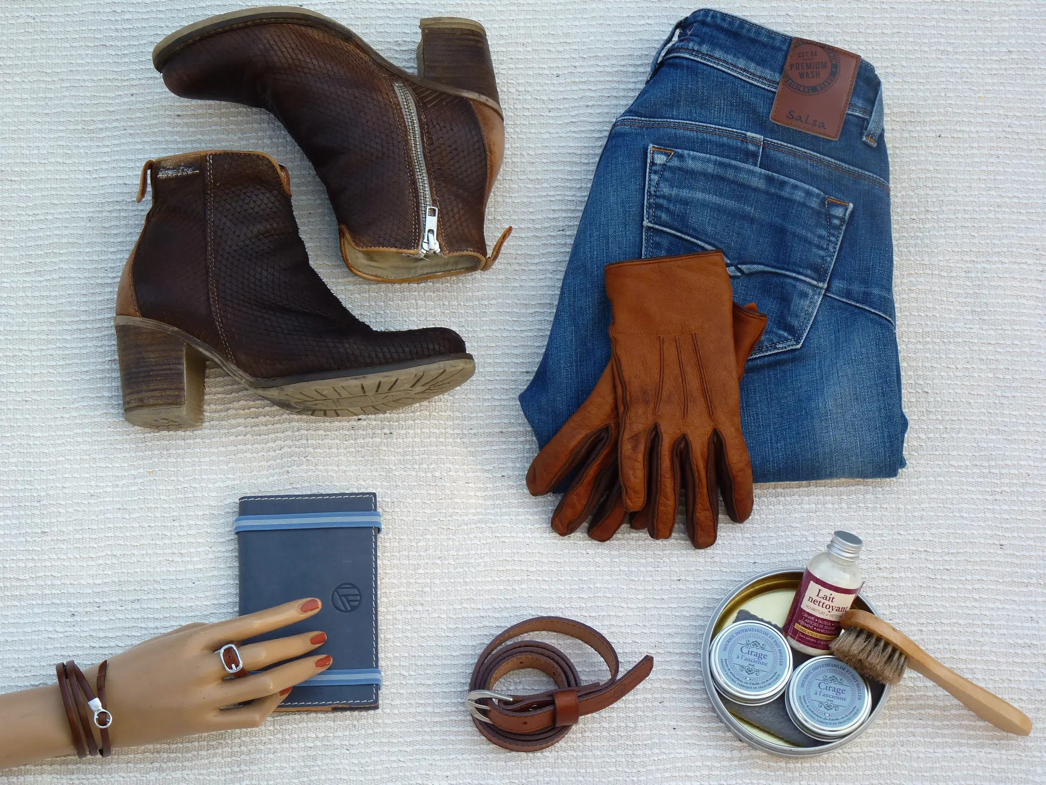 Bottes en cuir marron, jeans, gants en cuir, accessoires de mode et produits d'entretien sur fond clair.