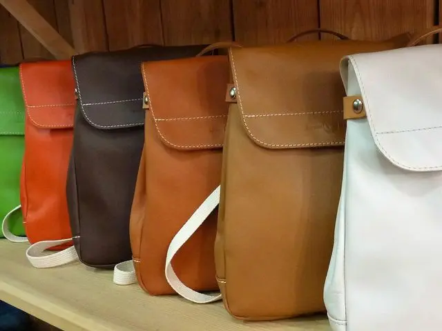 Cinq sacs à dos en cuir de différentes couleurs alignés sur une étagère en bois.